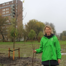 Vrouw met groene jas en boom in de hand. Op de achtergrond bomen en een flatgebouw.