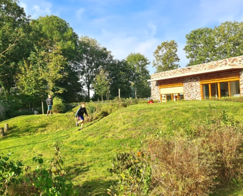Afbeelding van een groene heuvel met rechtsachter een gebouw en linksachter bomen. In het midden twee rennende kinderen